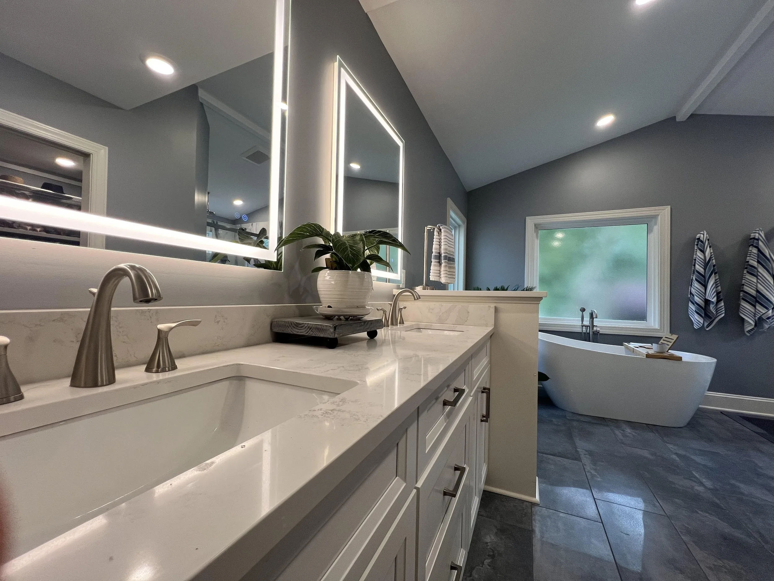 White double vanity with undermount sinks and lighted mirrors, and a freestanding tub under a frosted window beyond